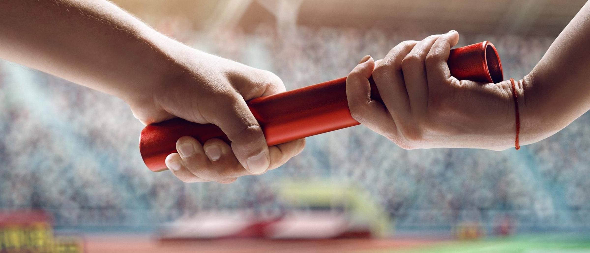 Two hands exchanging a red baton in a blurred stadium